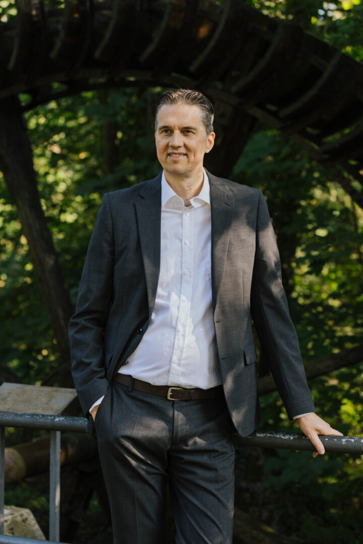 Gunnar Johannen steht im Freien, während er an ein Geländer gelehnt ist, trägt er einen dunkelblauen Anzug und ein helles Hemd.