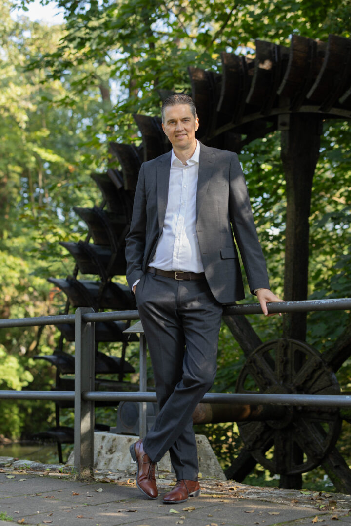 Gunnar Johannen steht im Freien und lehnt sich an ein Geländer an. Er trägt einen dunkelblauen Anzug und ein helles Hemd.