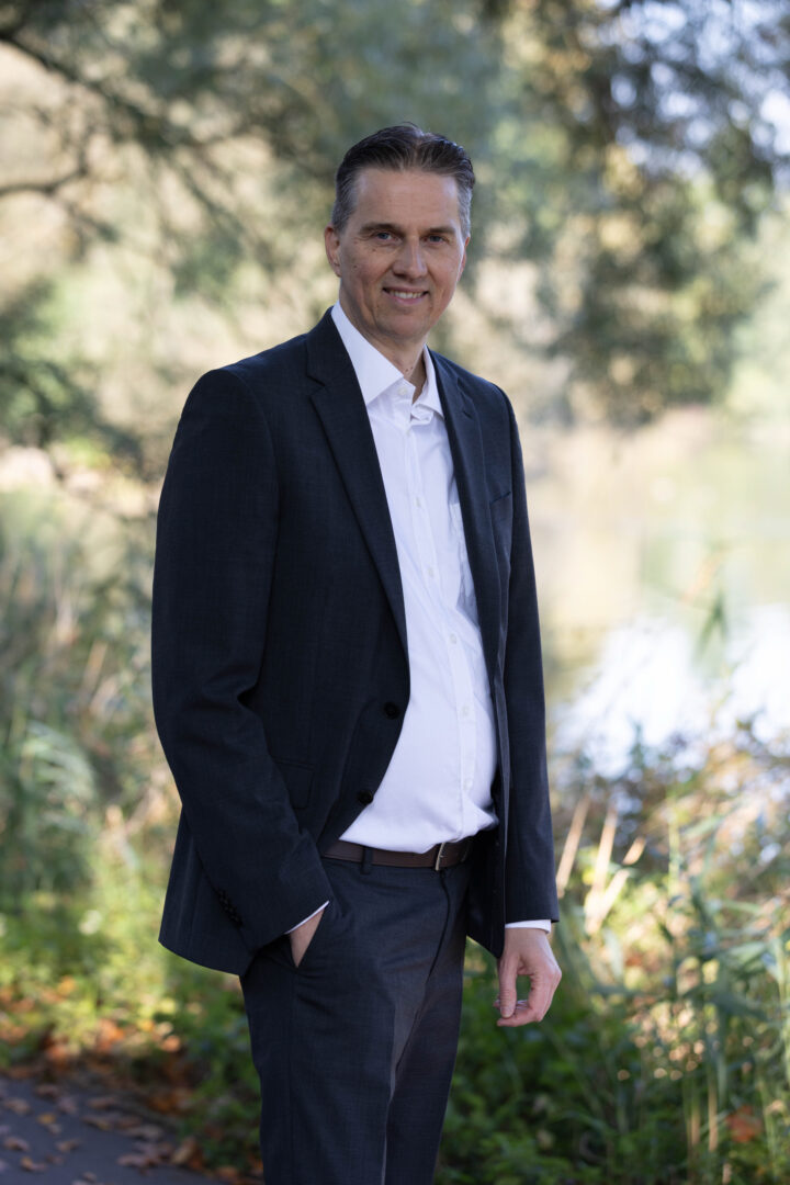 Gunnar Johannen steht in der Natur. Er trägt einen dunkelblauen Anzug und ein helles Hemd.