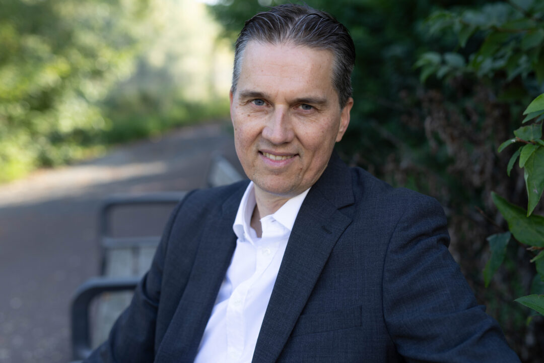 Gunnar Johannen sitzt im Freien auf einer Bank. Er trägt ein dunkelblaues Jackett und ein helles Hemd.