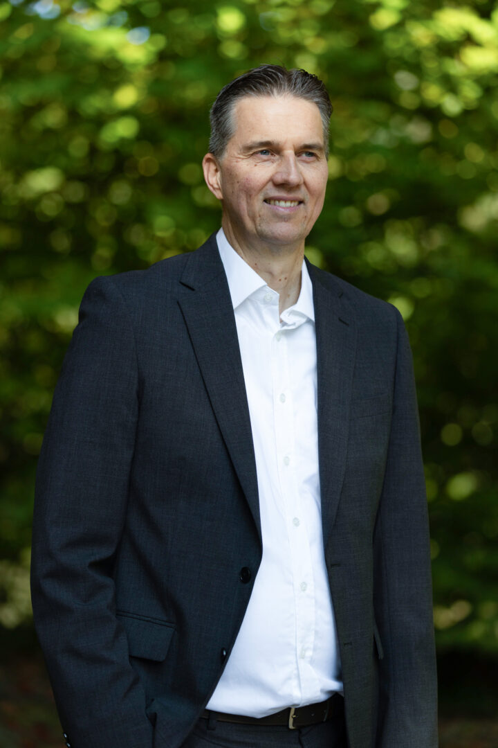 In der Natur steht Gunnar Johannen vor einem Gebüsch mit einem dunkelblauen Anzug und einem hellen Hemd.